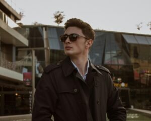 A fashionable man in sunglasses and a coat stands confidently in urban Ankara setting.